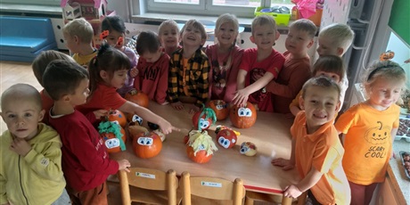 Powiększ grafikę: Grupa dzieci w pomarańczowych ubraniach w dzień dyni stoją uśmiechnięte przy stoliku na którym leżą ozdobionedynie.