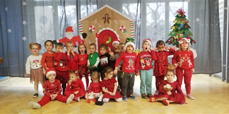 Powiększ grafikę: Dzieci ubrane na czerwono w czapkach mikołajowych na tle dekoracji Mikołajkowej.