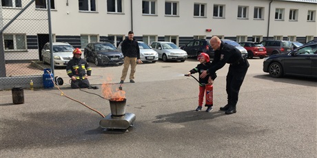 Powiększ grafikę: Dziecko z pomocą strażaka gasi płomień gaśnicą.